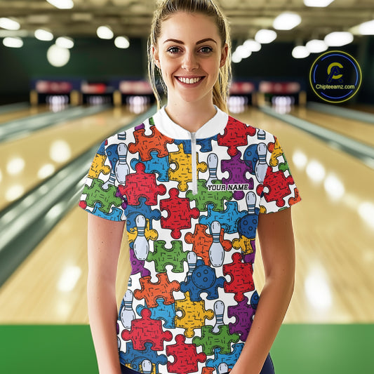 A woman stands in a bowling alley wearing a short-sleeve collared bowling shirt covered in colorful puzzle-piece graphics and bowling pin illustrations. The shirt has a white collar and a white chest area with the text 'YOUR NAME'.