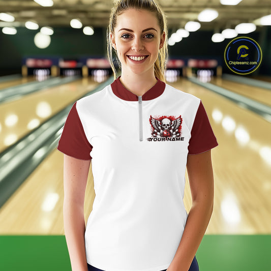 A woman stands in a bowling alley wearing a bowling shirt with quarter zip collar, red skull-themed bowling graphic with black wings, pins, bowling ball