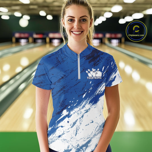 Front view of a female bowler in a bowling alley wearing a white and blue bowling quarter-zip shirt with a dynamic splatter jersey pattern