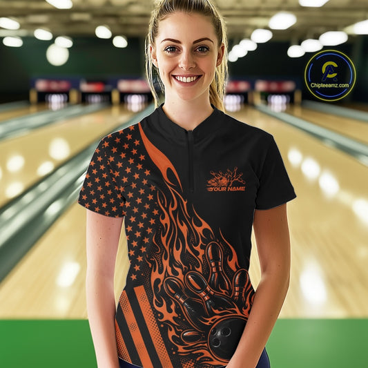 Female model at a bowling lane wearing a black bowling quarter-zip shirt with an orange flaming bowling ball hitting the pins and American flag artwork.