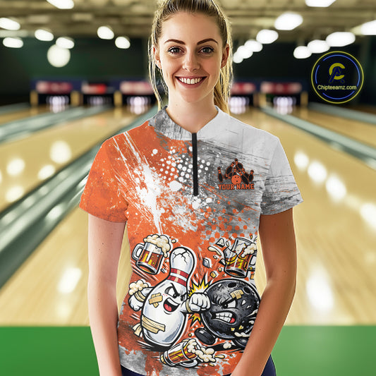 Front view of a female model in a bowling lane wearing an orange and white grunge splatter bowling quarter-zip shirt with funny cartoon bowling ball and pin fight scene.