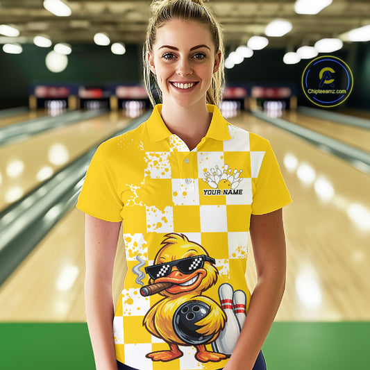 A woman stands in a bowling alley wearing a bright yellow and white checkered polo shirt with a cartoon duck graphic and the text 'YOUR NAME'. The background shows blurred bowling lanes, and a circular 'Chipleanz.