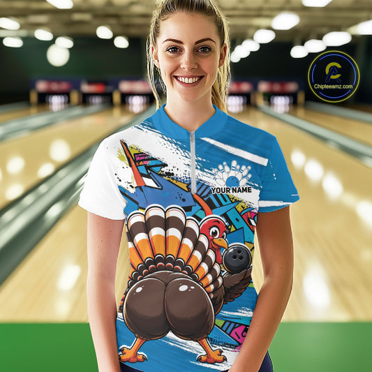 Front view of a happy female model in a bowling alley wearing a blue custom quarter zip bowling shirt with a funny graffiti turkey holding a bowling ball, paint splatter accents, and a personalized name.