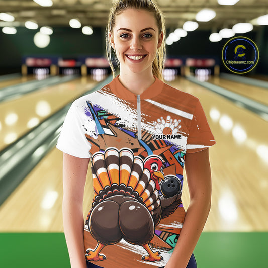 Front view of a happy female model in a bowling alley wearing an orange custom quarter zip bowling shirt with a funny graffiti turkey holding a bowling ball, paint splatter accents, and a personalized name.