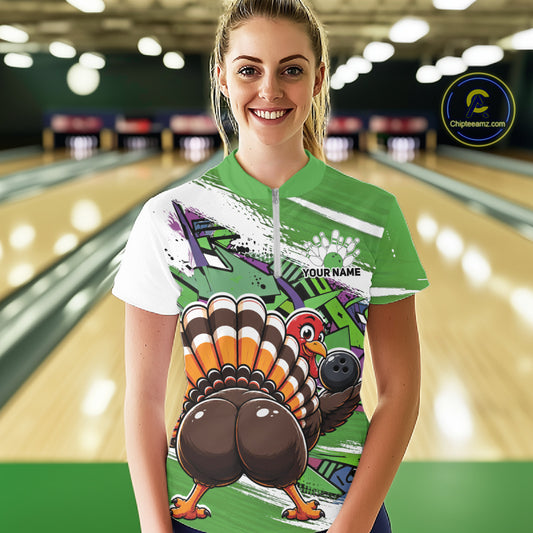 Front view of a happy female model in a bowling alley wearing a green custom quarter zip bowling shirt with a funny graffiti turkey holding a bowling ball, paint splatter accents, and a personalized name.