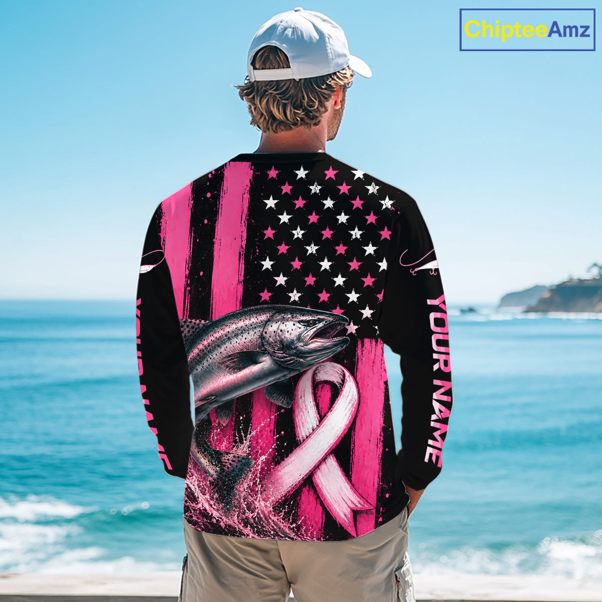 A man viewed from behind wearing a long-sleeve black shirt with a pink and white patriotic fish graphic, stars-and-stripes pattern, and pink ribbon design, standing by the ocean. The shirt includes the text 'YOUR NAME' on the sleeve.