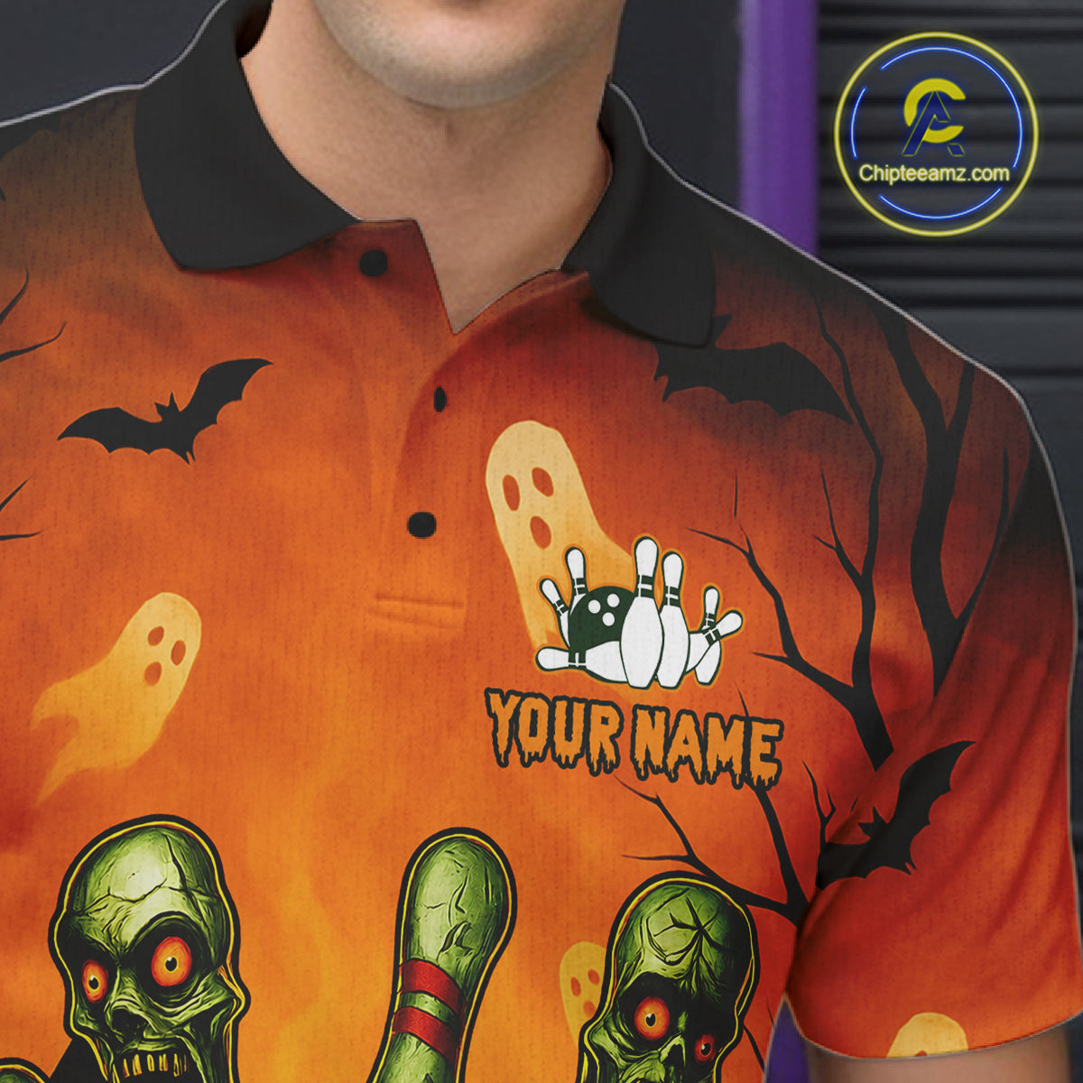 A close-up of a person wearing a short-sleeve polo shirt with a Halloween-themed graphic print. The shirt features an orange and black gradient background with illustrations of bats, ghosts, a zombie head, bowling pins, and the text 'YOUR NAME' with a custom bowling hand graphic above it.