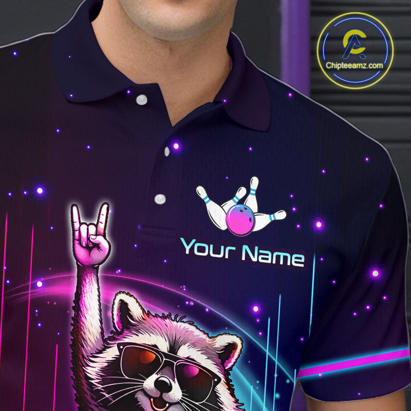 A close-up of a person wearing a dark navy polo shirt with a colorful custom graphic print. The print features a raccoon wearing sunglasses and making a rock-on gesture, along with bowling pins and the text 'Your Name' and a bowling ball.