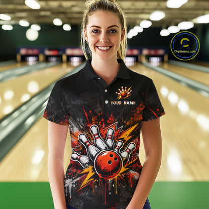 A woman stands in a bowling alley wearing a black short-sleeve polo shirt with a colorful bowling-themed graphic on the front. The shirt features a red bowling ball, white bowling pins, orange splatter effects, and the text 'YOUR NAME' on the chest.