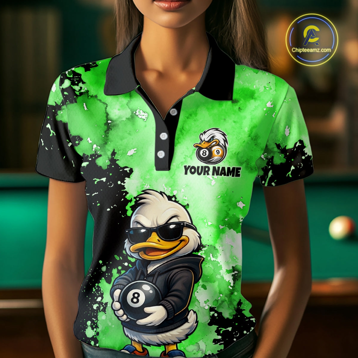 A woman wearing a short-sleeve polo shirt with a bright green and black splatter design, featuring a cartoon duck wearing sunglasses and holding an 8-ball, with the text 'YOUR NAME' and a bird-and-ball logo on the chest. The background is a blurred pool room.