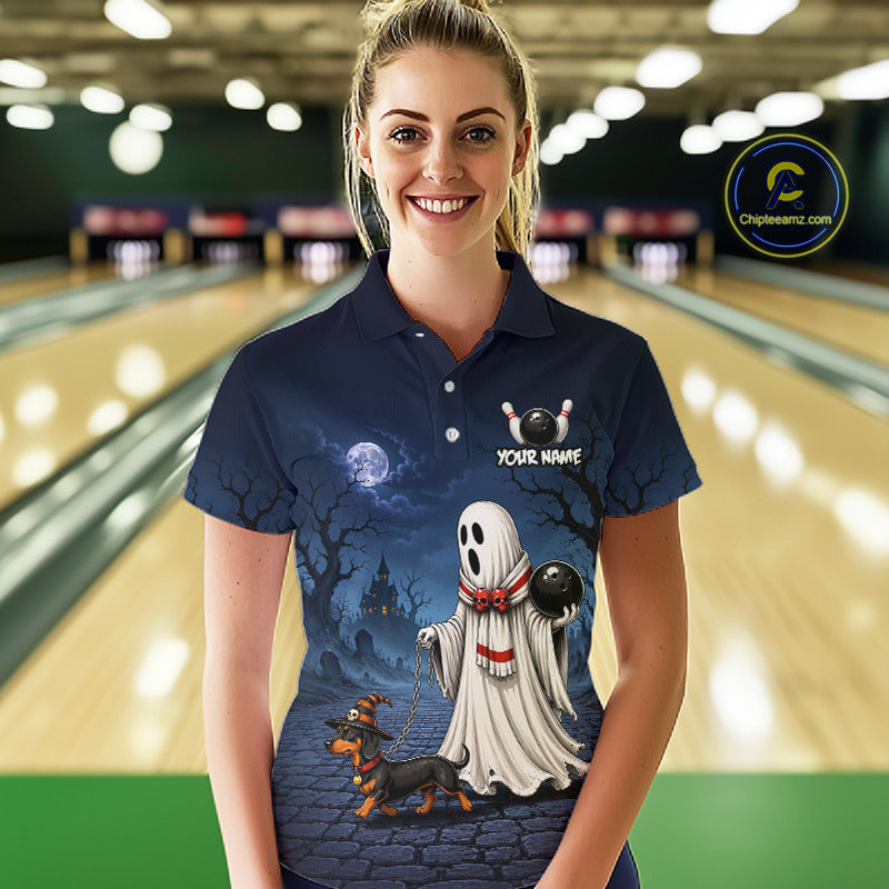 A woman wearing a navy blue custom polo shirt with a Halloween graphic print featuring a ghost, a dachshund, a cat, and a haunted house, set against a blurred bowling alley background.