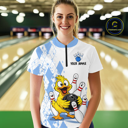 A woman stands in a bowling alley wearing a white short-sleeve bowling jersey with a blue collar and a large cartoon yellow duck holding bowling pins and a bowling ball on the front. The jersey has a blue and white argyle pattern on the sleeves and upper chest, a black zipper at the neck, and the text 'YOUR NAME' with a blue paw print above it.