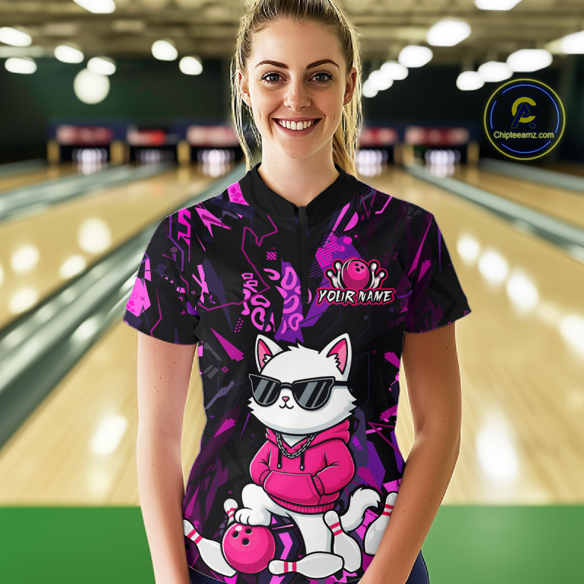 A woman standing in a bowling alley wearing a black short-sleeve bowling shirt with a colorful cartoon cat graphic and pink bowling balls. The background shows blurred bowling lanes and lights.