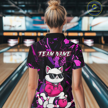 A woman viewed from behind wearing a short-sleeve bowling-themed polo shirt with a black and purple abstract background, a cartoon white cat wearing sunglasses and a pink hoodie, and the text 'TEAM NAME' across the upper back. Bowling lanes are visible in the blurred background.