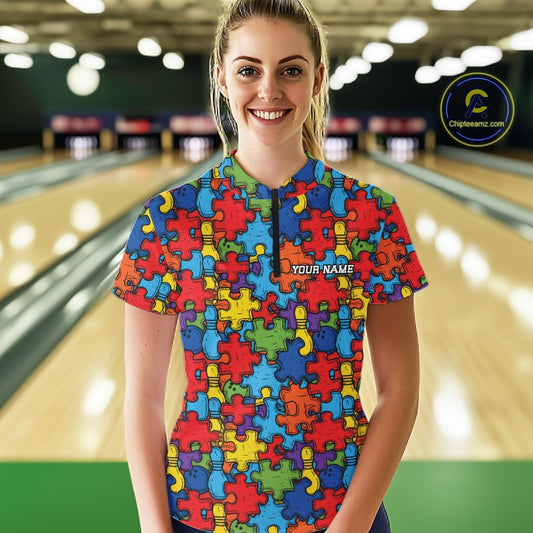 A woman standing in a bowling alley wearing a colorful short-sleeve quarter zip shirt with a puzzle-piece pattern. The shirt features a blue collar and a half-zip neckline with 'YOUR NAME' printed on the chest.
