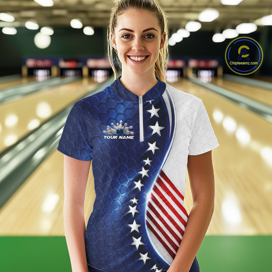 A woman stands in a bowling alley wearing a short-sleeve patriotic bowling shirt with a blue and white design, white stars, and red and white stripes. The shirt has a mock neck and a small logo on the left chest that reads 'YOUR NAME'.