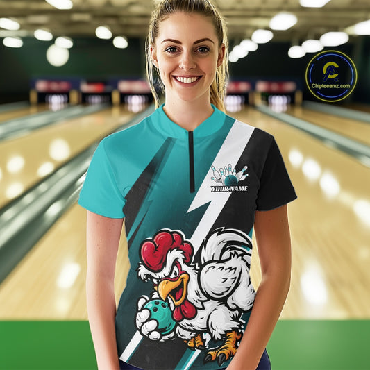 A woman standing in a bowling alley wearing a custom short-sleeve bowling jersey with a teal and black design, featuring a cartoon rooster mascot holding a bowling ball. The jersey has a mock neck and a black zipper at the collar.
