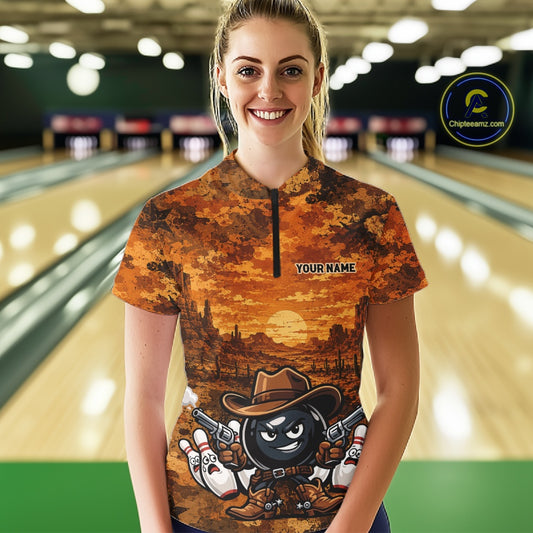 A woman stands in a bowling alley wearing a short-sleeve quarter-zip bowling shirt with a detailed western-themed graphic print featuring a cartoon bowling ball character. The shirt has a warm orange and brown sunset landscape design with the text 'YOUR NAME' on the chest.