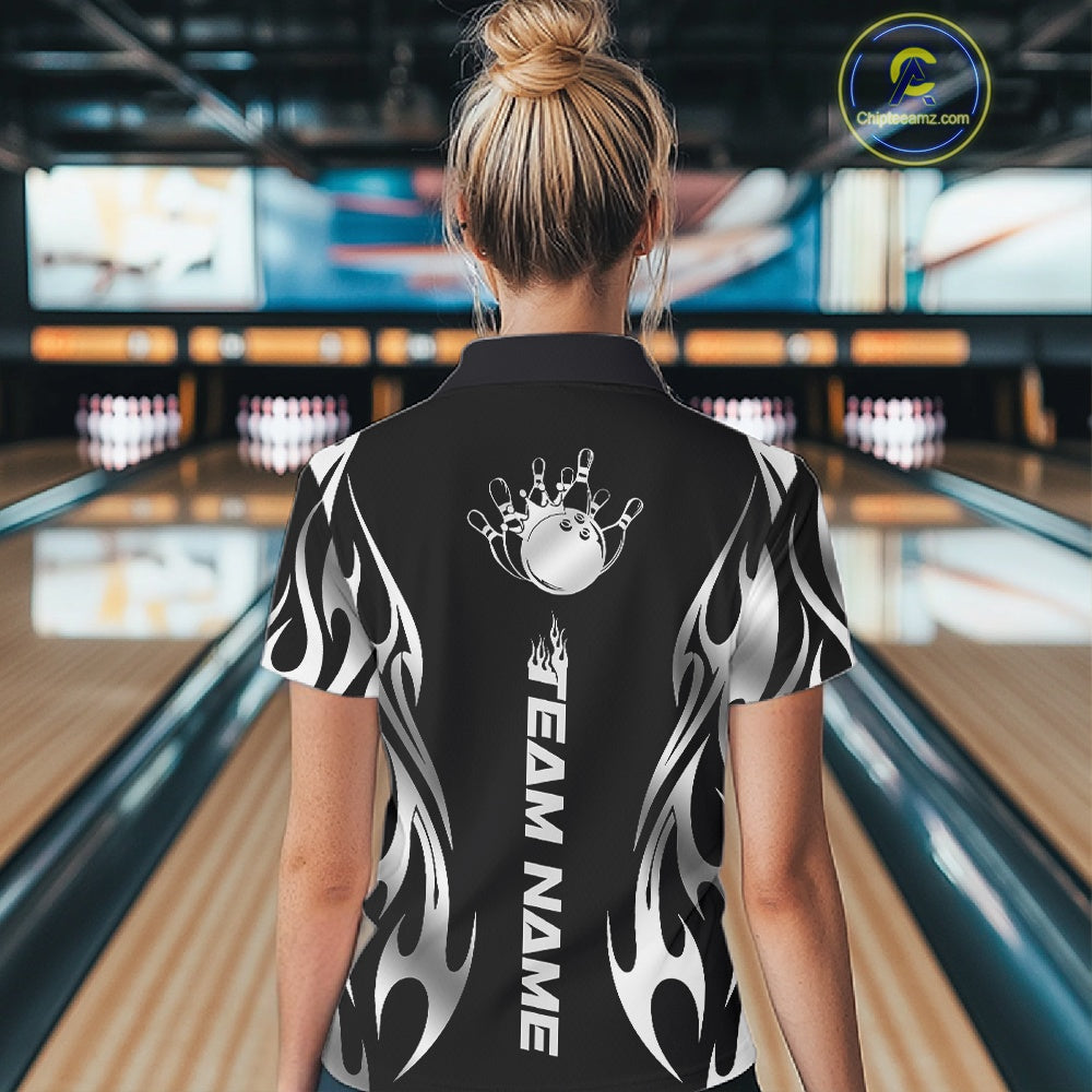Female model back view at a bowling lane wearing a black polo shirt featuring a silver metal tribal flame design with a bowling ball striking pins in dynamic motion.