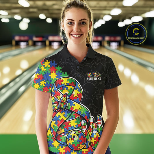 Female model in a bowling lane wearing a black polo shirt, front view showing a large autism awareness ribbon filled with puzzle pattern, colorful butterfly, and bowling ball striking pins artwork