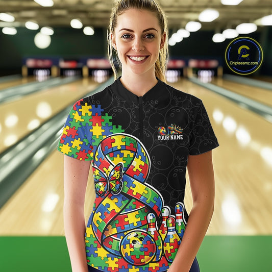 Female model in a bowling lane wearing a black quarter-zip shirt, autism ribbon with puzzle pattern, butterfly accent, and bowling ball striking pins artwork.
