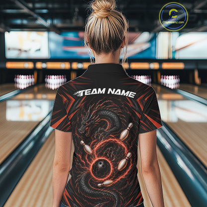 A woman viewed from behind wearing a black bowling-themed short-sleeve polo shirt with a orange and black dragon graphic and white 'TEAM NAME' text on the back, standing in a bowling alley.