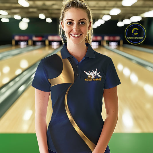 A woman stands in a bowling alley wearing a navy blue short-sleeve polo shirt with a gold abstract graphic and a white bowling pins logo with the text 'YOUR NAME'. 