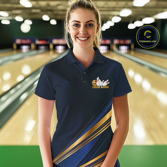 A woman stands in a bowling alley wearing a navy blue short-sleeve polo shirt with gold diagonal stripes and a bowling-themed graphic on the left chest. The background shows blurred bowling lanes with bright overhead lights.