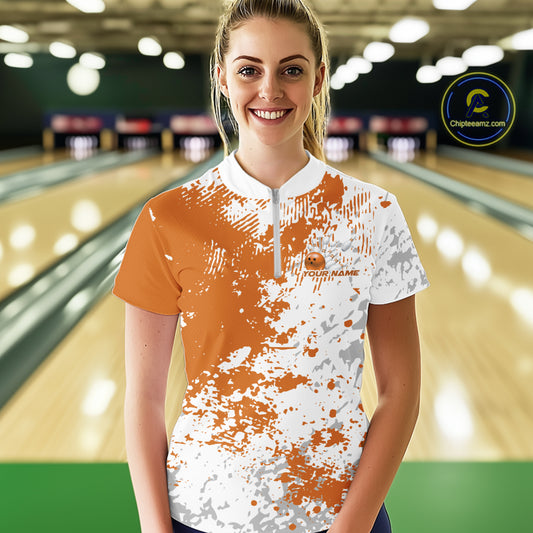 Front view of a female model in a bowling lane wearing a bowling quarter-zip shirt with orange and white splatter background and dynamic athletic styling.