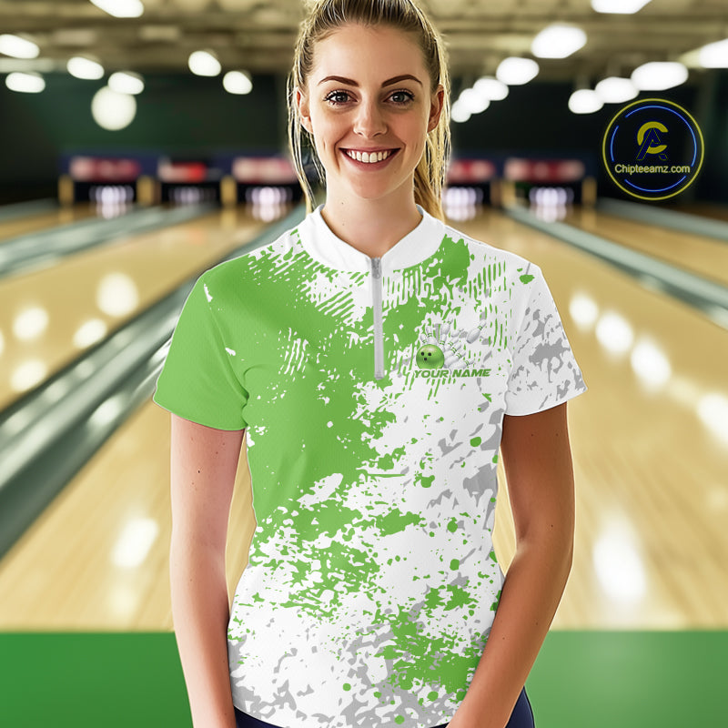 Front view of a female model in a bowling lane wearing a green and white splatter bowling quarter-zip shirt with minimalist bowling-themed design accents.