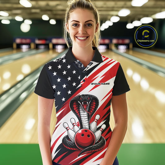 A woman stands in a bowling alley wearing a black, white, and red bowling shirt with stars, diagonal stripes, a cobra, pins, and an red ball.