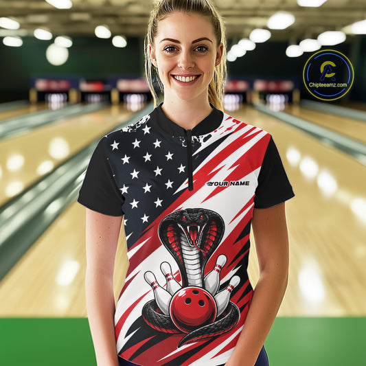 A woman stands in a bowling alley wearing a black, white, and red bowling shirt with stars, diagonal stripes, a cobra, pins, and an red ball.