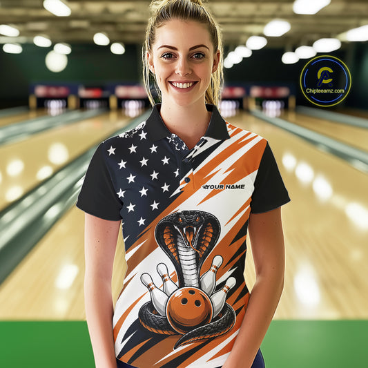 A woman stands in a bowling alley wearing a black, white, and orange bowling shirt with stars, diagonal stripes, a cobra, pins, and an orange ball.