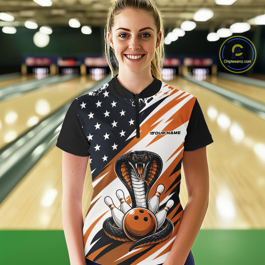 A woman stands in a bowling alley wearing a black, white, and orange bowling shirt with stars, diagonal stripes, a cobra, pins, and an orange ball.