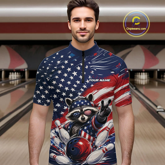 Front view of a male model at a bowling lane wearing a bowling quarter-zip shirt with a patriotic raccoon flashing a peace sign as a red, white, and blue bowling ball crashes through falling pins beneath a flowing American flag.