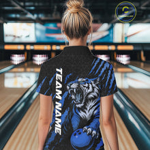 Load image into Gallery viewer, Back view of a female model wearing a black and mid blue bowling polo shirt in a bowling lane, featuring a roaring tiger holding a mid blue bowling ball with coordinated claw-stripe graphics and a geometric background pattern.