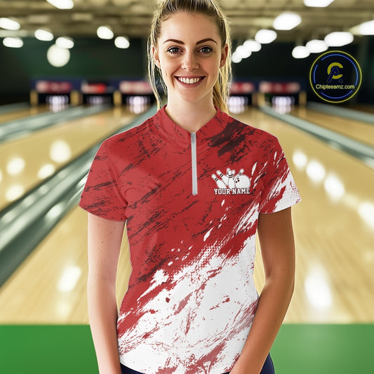 Front view of a female model in a bowling alley wearing a bowling quarter-zip shirt featuring a white paint splatter distressed sportswear design on a red shirt.
