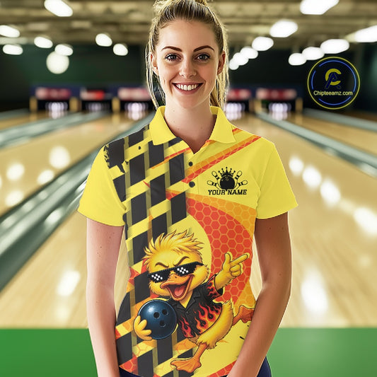 A woman stands in a bowling alley wearing a bright yellow custom-printed polo shirt with a cartoon duck graphic, black and yellow checkered pattern, and a black logo that reads 'YOUR NAME'. Bowling lanes are visible in the blurred background.
