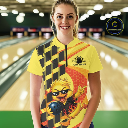 A woman stands in a bowling alley wearing a bright yellow short-sleeve bowling shirt with a black-and-yellow checkerboard pattern on one side, a red honeycomb graphic on the other, and a cartoon duck character holding a bowling ball. The shirt has a black zipper at the collar and a black chest graphic that reads 'YOUR NAME' with a bowling pin icon above it.