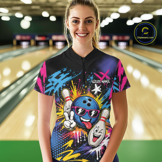 A woman stands in a bowling alley wearing a short-sleeve black quarter zip shirt with a colorful bowling-themed graphic print featuring cartoon bowling balls and pins, pink and blue paint splatters