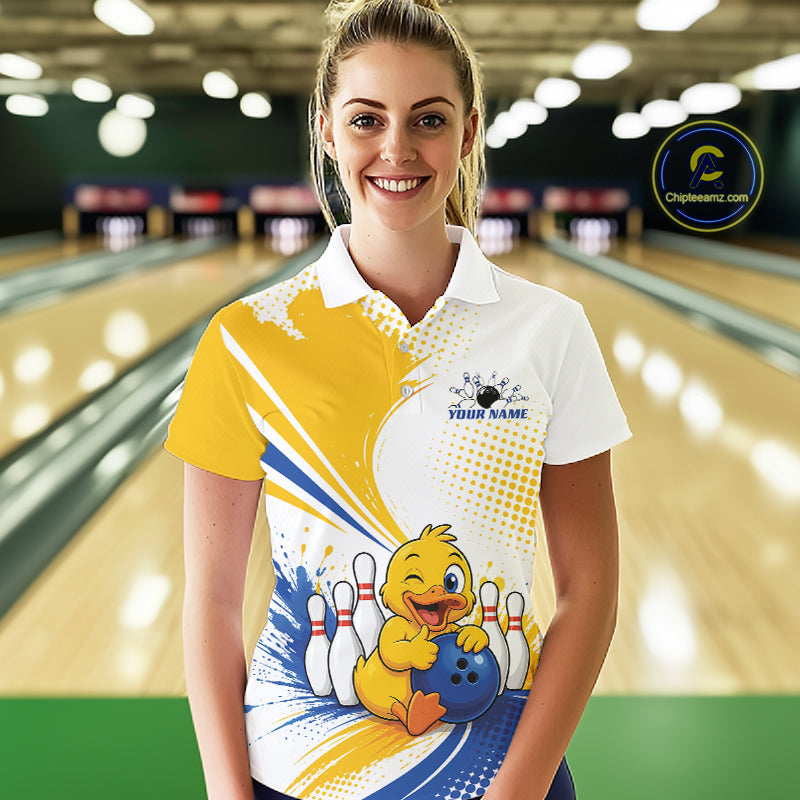 A woman stands in a bowling alley wearing a custom printed short-sleeve polo shirt. The shirt features a white collar and upper section, with a yellow, blue, and white graphic design on the lower half showing a cartoon duck holding a bowling ball.