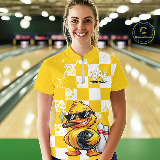 A woman stands in a bowling alley wearing a bright yellow and white checkered short-sleeve bowling shirt with a cartoon duck graphic and a custom name printed on the chest. A circular 'Chilewich.