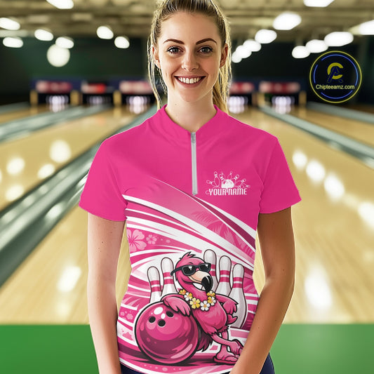 Female model at a bowling lane wearing a pink bowling quarter-zip shirt with a tropical flamingo in sunglasses beside a bowling ball.