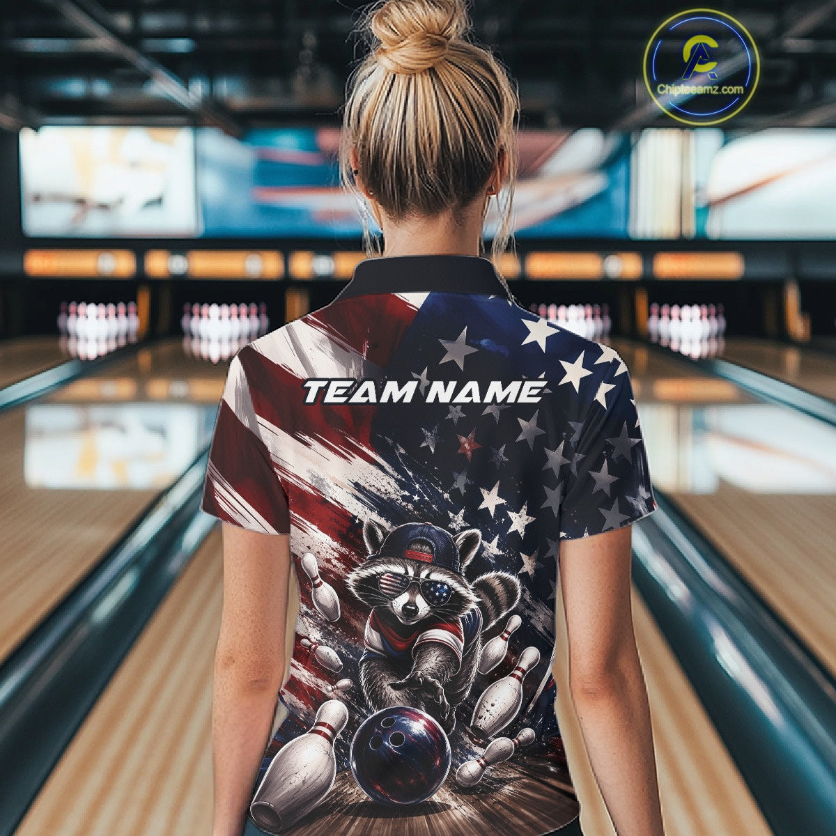 Back view of a female model inside a bowling center wearing a bowling quarter-zip shirt with a raccoon in patriotic sunglasses rolling a bowling ball through scattered pins against a distressed American flag backdrop.