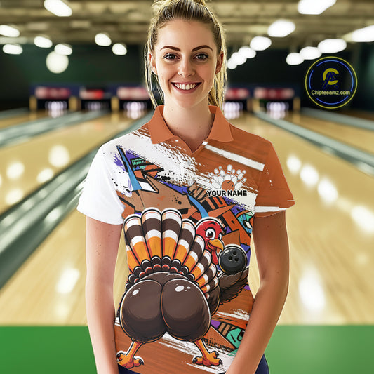Front view of a female model wearing an orange custom bowling polo shirt with a funny graffiti turkey holding a bowling ball, paint splatter accents, and a personalized name in a bowling alley.