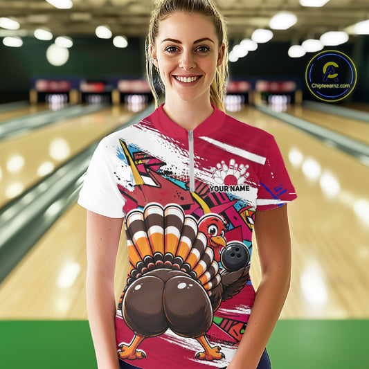 Front view of a happy female model in a bowling alley wearing a red custom quarter zip bowling shirt with a funny graffiti turkey holding a bowling ball, paint splatter accents, and a personalized name.