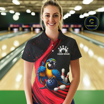 A woman stands in a bowling alley wearing a black short-sleeve polo shirt with a red graphic design, a blue and yellow parrot, bowling pins, and a blue bowling ball. The shirt also features a white bowling pin graphic and the text 'YOUR NAME' on the chest.