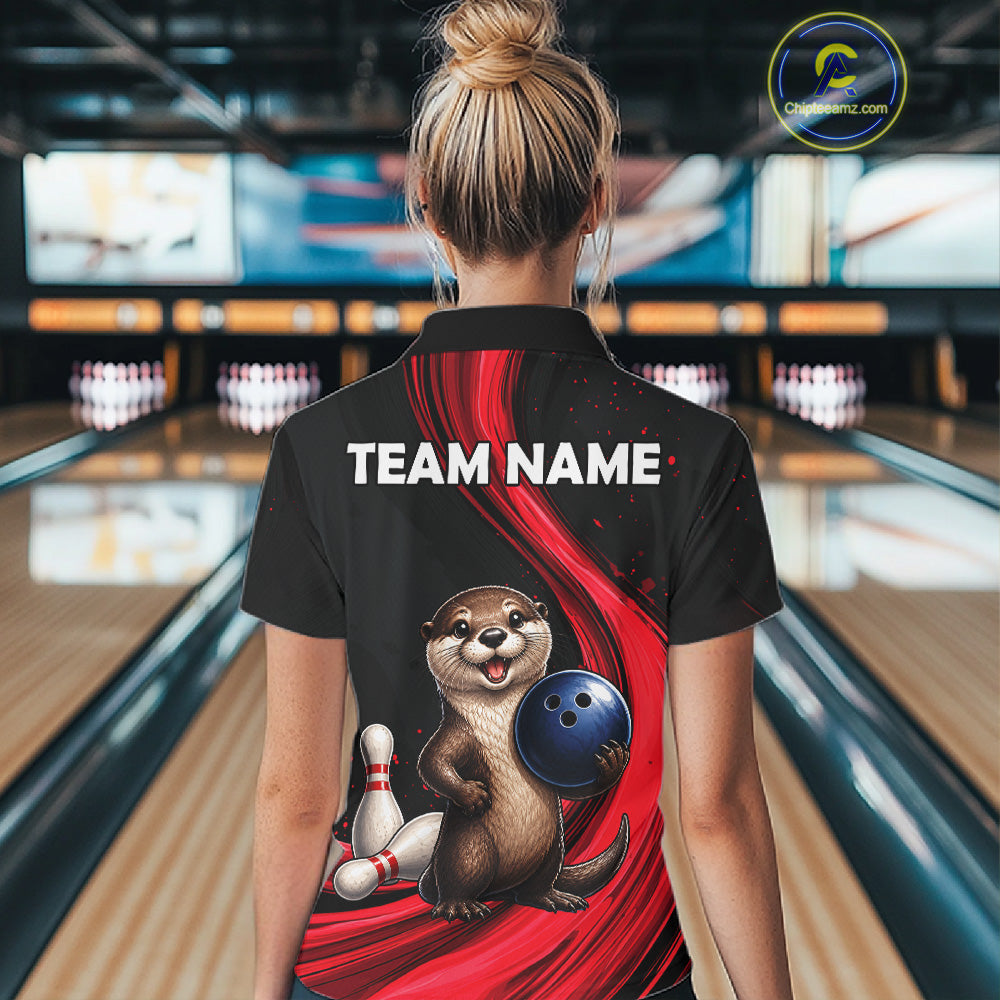 A woman viewed from behind wearing a black short-sleeve bowling-themed polo shirt with a large cartoon otter graphic, red swoosh design, and the text 'TEAM NAME' on the back. Bowling lanes are visible in the blurred background.