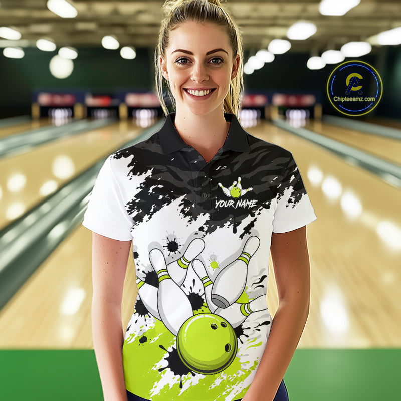 A woman standing in a bowling alley wearing a custom bowling-themed polo shirt. The shirt features a black and white paint splatter design with a graphic of bowling pins, bowling balls, and a bowling pin collision, along with the text 'YOUR NAME' and a bowling pin logo.