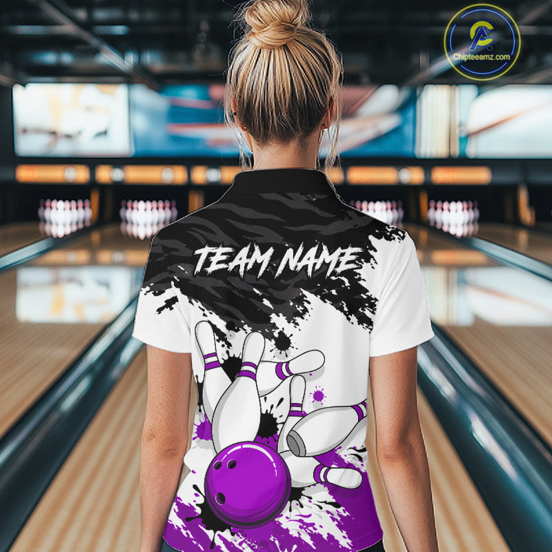 A woman viewed from behind wearing a customized bowling-themed polo shirt in a bowling alley. The back of the white and black polo shirt features a purple bowling ball and pins graphic, with 'TEAM NAME' printed in white text.